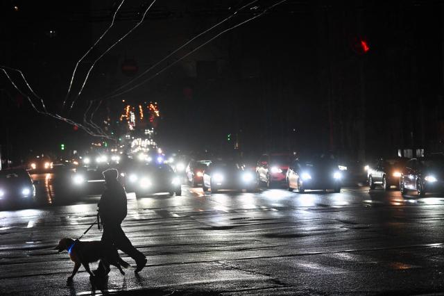 A man carries a dog as they cross a street in front of vehicles with headlights driving down a non-illuminated street during a power outage in Kyiv on December 27, 2025, following Russian missile and drone attacks on Ukrainian energy infrastructure amid the Russian invasion of Ukraine. (Photo by Sergei SUPINSKY / AFP)