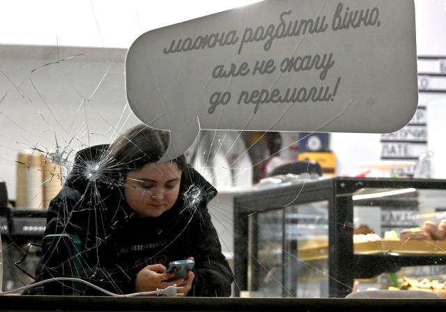 A girl looks at smartphone as she sits at cafe's shopwindow broken by fragments and decorated with placard reading "You can break a window, but not our thirst for victory", in Kyiv on December 27, 2025, following Russian missile and drone attacks amid the Russian invasion of Ukraine. (Photo by Sergei SUPINSKY / AFP)