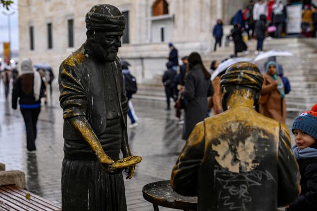 People walk past the statue representing a seller of Turkish bagels "Simit" at the New Mosque Square, in the Eminonu district of Istanbul, on December 27, 2025. Simit roams the streets of Turkey as surely as cats. But the price of this small, round, ring-shaped bread, studded with sesame seeds, is also a stark indicator of the dizzying price increases. (Photo by Yasin AKGUL / AFP)