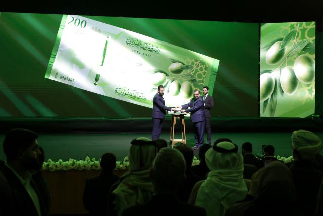 Syria's President Ahmed al-Sharaa (L) and Syria's Central bank chief Abdul Qadir al-Hasriya (R) hold one of the country's new revamped currency notes at the Conference Palace in the Syrian capital Damascus on December 29, 2025. Syria's central bank chief said on December 25 that the country's currency would start to be replaced with a revamped version from January 1, as Damascus tries to relaunch its economy after emerging from the rule of ousted leader Bashar al-Assad last year. (Photo by Bakr ALkasem / AFP)