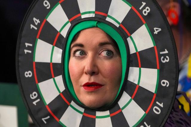 A woman wearing dartboard-themed headgear watches during the World Darts Championship at Alexandra Palace in London on December 28, 2025. Dressed as Elvis, Asterix or Snow White, usually with a drink in hand, fans transform London's Alexandra Palace into an overheated darts temple for a world championship that resembles a Christmas carnival. Some are there for the darts, but for others the annual event is about dressing up, having a drink and soaking up the raucous atmosphere. (Photo by CARLOS JASSO / AFP)