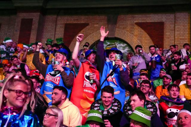 Spectators dressed up as Pringles cheer during the World Darts Championship at Alexandra Palace in London on December 28, 2025. Dressed as Elvis, Asterix or Snow White, usually with a drink in hand, fans transform London's Alexandra Palace into an overheated darts temple for a world championship that resembles a Christmas carnival. Some are there for the darts, but for others the annual event is about dressing up, having a drink and soaking up the raucous atmosphere. (Photo by CARLOS JASSO / AFP)