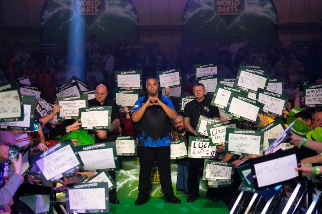 Netherlands' Jermaine Wattimena (C) poses with supporters as he walks on for his match against Scotlands Gary Anderson during the World Darts Championship at Alexandra Palace in London on December 28, 2025. Dressed as Elvis, Asterix or Snow White, usually with a drink in hand, fans transform London's Alexandra Palace into an overheated darts temple for a world championship that resembles a Christmas carnival. Some are there for the darts, but for others the annual event is about dressing up, having a drink and soaking up the raucous atmosphere. (Photo by CARLOS JASSO / AFP)