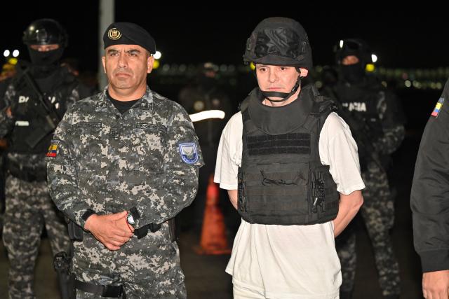 Roberto Alvarez (R), alias "El Gerente," an Ecuadorean alleged leader of a Colombian guerrilla group, is escorted by the Commander-in-Chief of Ecuador's National Police, Pablo Davila (L) and Ecuador's Interior Minister John Reimberg (out of frame), upon landing at the Police base in Jose Joaquin de Olmedo International Airport in Guayaquil, Ecuador, on December 29, 2025, after being transferred from the United Arab Emirates. Alvarez, wanted by Ecuadorean authorities for the killing of 11 soldiers, has been identified by the government as the head of the Border Commands, a dissident faction of the now-defunct Colombian guerrilla group FARC that did not adhere to the 2016 peace agreement. (Photo by Marcos PIN / AFP)