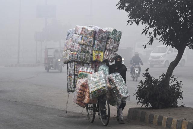 A vendor with his face covered in cloth carries food items on a bicycle along a street amid dense smog in Lahore on December 30, 2025. (Photo by Arif ALI / AFP)