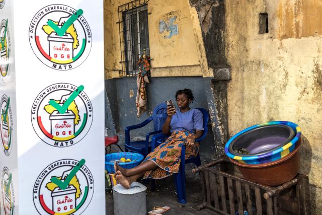 A Guinean woman holds and looks at her smartphone next to a logo of the General Directorate of Elections (DGE) near its offices during the announcement of official partial results of Guinea’s presidential election in Conakry, on December 30, 2025. Guinea's junta chief, who had pledged not to run for office after seizing power four years ago, has a large lead in presidential elections held at the weekend, according to initial results published late December 29. (Photo by PATRICK MEINHARDT / AFP)