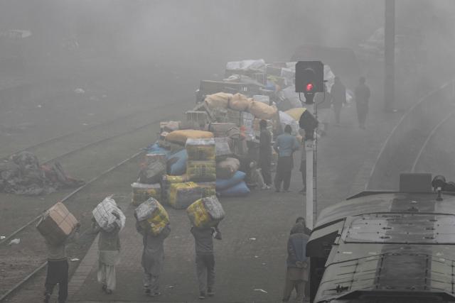 Workers carry goods as people wait for their train at a railway station amid dense fog on a cold winter morning in Lahore on January 2, 2026. (Photo by Arif ALI / AFP)