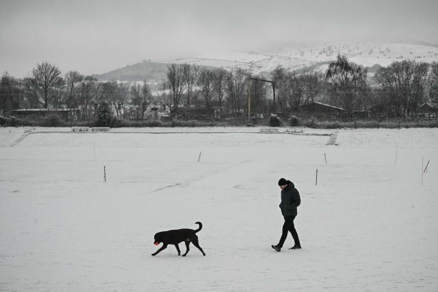 A man walks his dog across a snow-covered cricket pitch in the town of Glossop, Derbyshire, northern England on January 6, 2026, after a light snow covered the region overnight. The UK's Met Office issued fresh weather warnings for January 5-6 for snow and ice for Scotland, Northern Ireland and parts of northern England and said cold weather health alerts for all English regions would remain in place until January 9. (Photo by Oli SCARFF / AFP)