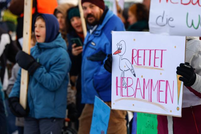 Midwifes and family members protest with placards against the "Hebammenhilfe-Vertrag" (Midwifery Care Agreement) on the sidelines of the CSU party faction retreat at Seeon Abbey in Seeon, southern Germany, on January 6, 2026. A controversial new midwifery care agreement entered into force on November 1, 2025 and sparks fears that many midwives could stop exercising. (Photo by Michaela STACHE / AFP)