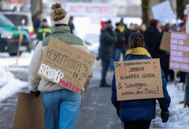Midwifes and family members protest with placards against the "Hebammenhilfe-Vertrag" (Midwifery Care Agreement) on the sidelines of the CSU party faction retreat at Seeon Abbey in Seeon, southern Germany, on January 6, 2026. A controversial new midwifery care agreement entered into force on November 1, 2025 and sparks fears that many midwives could stop exercising. (Photo by Michaela STACHE / AFP)