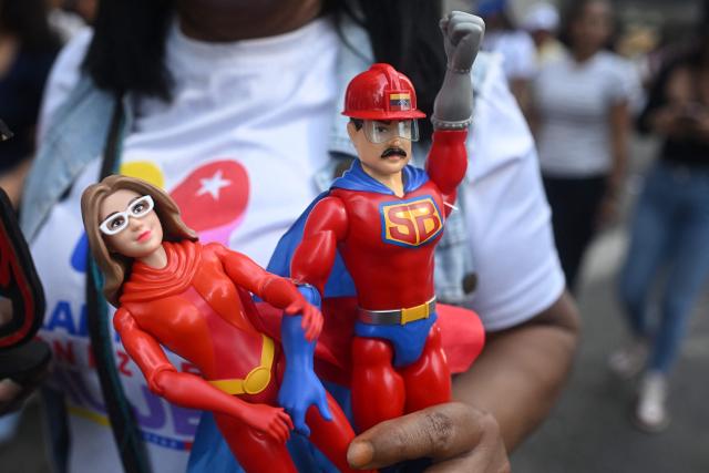 A woman holds toys of the caped superhero named "Super-Bigote" (Super-Mustache) and "Cilita," as well as a drawing of Venezuela's independence hero Simon Bolivar during a rally in support of ousted Venezuela's President Nicolas Maduro and his wife Cilia Flores in Caracas on January 6, 2026. US forces killed 55 Venezuelan and Cuban military personnel during their stunning raid to capture Nicolas Maduro, tolls published by Caracas and Havana showed on January 6. (Photo by Federico PARRA / AFP)