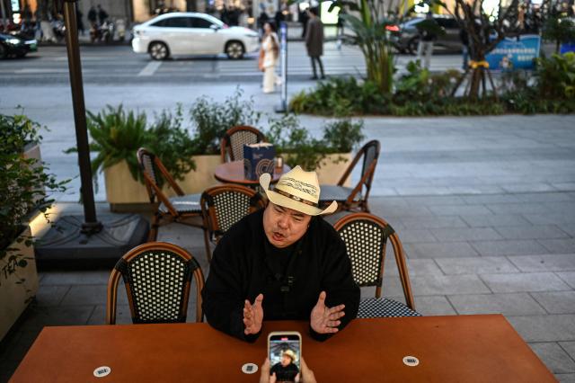 The photo taken on December 15, 2025 shows Donald Trump's impersonator Ryan Chen speaking as his assistant records a video at a milk tea shop in southwestern China's Chongqing municipality. Outstretching his hands in a signature Donald Trump pose, impersonator Ryan Chen mimics the US president's voice and gestures with such accuracy he has become a social media phenomenon with his funny videos. (Photo by Jade GAO / AFP) / TO GO WITH: China-US-social-comedy-Trump, PROFILE by Ludovic EHRET