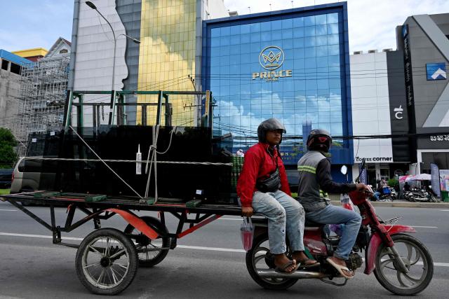 (FILES) Workers ride their motor-cart loaded past a branch of the Prince Bank in Phnom Penh on October 15, 2025. A Cambodian bank founded by accused scam boss Chen Zhi, who has been indicted by the United States and extradited to China, was ordered liquidated on January 8, 2026, Cambodia's central bank said. (Photo by TANG CHHIN Sothy / AFP)