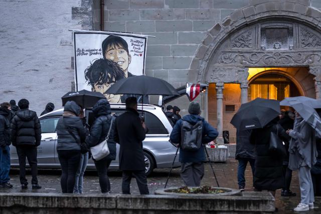 People stand next to a hearse and a banner depicting Arthur Brodard and his brother Benjamin at the end of the funeral of 16 year old Arthur Brodard who died in Crans-Montana, in Lutry on January 8, 2026. Fourty people, most of them teenagers, were killed while 116 were injured in a fire that ripped through a bar in the luxury Alpine ski resort of Crans-Montana on New Year's Eve. (Photo by Fabrice COFFRINI / AFP)