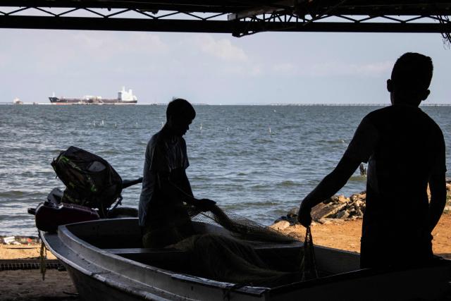 TOPSHOT - Two silhouettes of kids carrying a net at a fishing pier are seen in a picture, while in the background a crude oil tanker is docking in the Maracaibo Lake in Maracaibo, Venezuela, on January 8, 2026. On January 8, three of 11 vessels chartered by Chevron were transporting oil from Venezuela to the US, according to an AFP analysis of ship-tracking data, as export sanctions raised concerns over the South American country's storage capacity. (Photo by Maryorin Mendez / AFP)