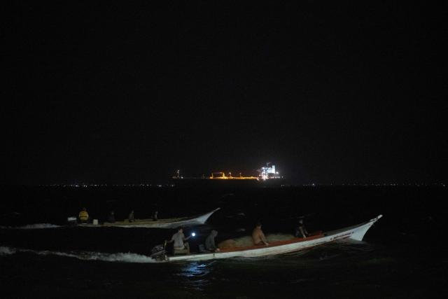 Two boats sail while in the background is seen a crude oil tanker docking in the Maracaibo Lake in Maracaibo, Venezuela, on January 9, 2026. On January 8, three of 11 vessels chartered by Chevron were transporting oil from Venezuela to the US, according to an AFP analysis of ship-tracking data, as export sanctions raised concerns over the South American country's storage capacity. (Photo by Maryorin Mendez / AFP)