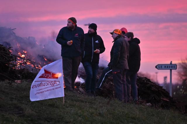 French farmers stand near a French agricultural union Jeunes agriculteurs (JA) flag at a roundabout as part of national protests against the EU-Mercosur agreement and the government's handling of the contagious nodular dermatitis (CND) epidemic, on the port of Le Havre, northwestern France on January 11, 2026. Around a hundred farmers spent the night at the entrance to the port of Le Havre, western France to set up a "filtering roadblock" on January 11, 2026 and check trucks in order to denounce the agreement signed by the EU with the Mercosur countries, a spokesperson told AFP. (Photo by Lou BENOIST / AFP)