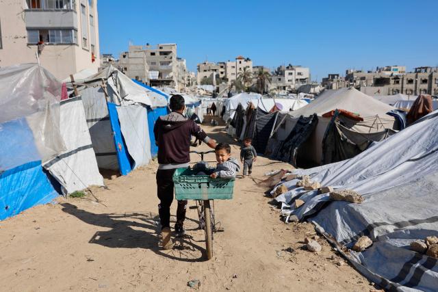 TOPSHOT - A child sits in a basket attached to a bicycle as a man pushes it past tent shelters housing displaced Palestinian families, in Gaza City on January 11, 2026. The majority of Gaza's 2.4 million people have been displaced, often multiple times, by the war that began with Hamas's attack on southern Israel on October 7, 2023. With displaced families living in tented camps, a serious concerns has been raised over their living conditions. (Photo by Omar AL-QATTAA / AFP)