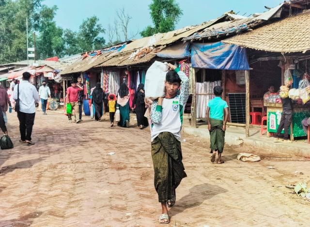 A Rohingya refugee carrying a sack walks across a market at the Kutupalong refugee camp in Ukhia on January 12, 2026. In Bangladesh's sprawling Rohingya camps of Cox's Bazar, where more than a million refugees forced to flee Myanmar live in squalid conditions, hope is a fragile but persistent force. In rows of bamboo shelters and muddy lanes, refugees who escaped Myanmar nearly a decade ago await developments more than 8,000 kilometres (5,000 miles) away at the International Court of Justice (ICJ) in The Hague, where a genocide case against Myanmar opens on January 12. (Photo by MH Mustafa / AFP)