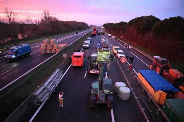 Farmers stand beside agricultural vehicles and bales of hay on the A64 motorway during a farmers' blockade to protest against the government's mandatory culling protocol for cattle herds affected by lumpy skin disease (dermatose nodulaire contagieuse) and the EU-Mercosur trade deal, in Carbonne, southwestern France on January 13, 2026. A roadblock that farmers have been maintaining for a month on the A64 motorway near Toulouse was being dismantled in the morning of January 13, 2026 by law enforcement, according to multiple sources. (Photo by Lionel BONAVENTURE / AFP)