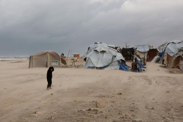 A young Palestinian shields himself from blowing sand next to tent shelters housing displaced families set up along the shore in Gaza City, as strong winter winds sweep the Palestinian enclave on January 13, 2026. A fragile ceasefire has been in place since October, following a deadly war waged by Israel in response to Hamas's unprecedented October 7, 2023 attack on Israel. Nearly 80 percent of buildings in Gaza have been destroyed or damaged by the war, according to UN data. (Photo by Omar AL-QATTAA / AFP)