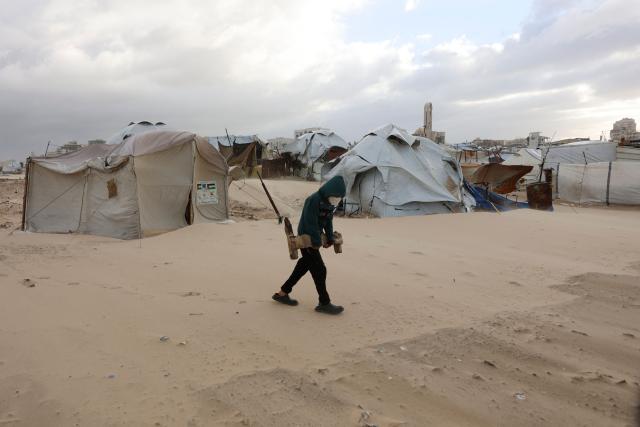 A young Palestinian carries a piece of wood past tent shelters housing displaced families set up along the shore in Gaza City as strong winter winds sweep the Palestinian enclave on January 13, 2026. A fragile ceasefire has been in place since October, following a deadly war waged by Israel in response to Hamas's unprecedented October 7, 2023 attack on Israel. Nearly 80 percent of buildings in Gaza have been destroyed or damaged by the war, according to UN data. (Photo by Omar AL-QATTAA / AFP)