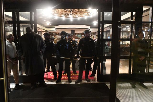 Local law enforcement stand at the entrance of the Graduate Hotel, where federal immigration agents are believed to be staying, while anti-ICE protesters demonstrate outside the hotel in Minneapolis on January 13, 2026. Hundreds more federal agents were heading to Minneapolis, the US homeland security chief said on January 11, brushing aside demands by the Midwestern city's Democratic leaders to leave after an immigration officer fatally shot a woman protester. In multiple TV interviews, US Homeland Secretary Kristi Noem defended the actions of the officer who shot and killed 37-year-old Renee Nicole Good, whose death has sparked renewed protests nationwide against President Donald Trump's immigration crackdown. (Photo by Octavio JONES / AFP)