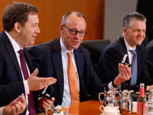 German Chancellor Friedrich Merz (C) sits between German Finance Minister and Vice Chancellor Lars Klingbeil and Chief of Staff and Minister for Special Tasks Thorsten Frei as he shows a 2-Euro silver coin commemorating former and late German Chancellor Konrad Adenauer that was distributed to the cabinet members at the start of the weekly cabinet meeting on January 14, 2026 at the Chancellery in Berlin. The coin was officially released on January 8, 2026 on the occasion of the 150th birthday of late German Chancellor Konrad Adenauer (Photo by Odd ANDERSEN / AFP)