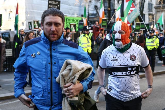Kneecap member DJ Provaí (R) and Kneecap manager Dan Lambert (L) walk past supporters and protesters (behind) as they arrive outside the Royal Courts of Justice ahead of the arrival of band member Liam O'Hanna, who performs under the stage name Mo Chara, in London on January 14, 2026. The singer Mo Chara is back before the courts on January 14 as British prosecutors challenge a decision to drop a terrorism charge against him. (Photo by Henry NICHOLLS / AFP)