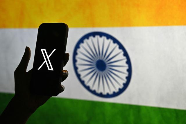 This photo illustration shows a person holding a smartphone bearing the logo of X, formerly known as Twitter, in front of the Indian national flag in Bengaluru on January 15, 2026. Elon Musk's platform X on January 14 announced measures to prevent its AI chatbot Grok from undressing images of real people, following global backlash over its generation of sexualized photos of women and children. India has said X removed thousands of posts and hundreds of user accounts in response to its complaints. (Photo by Idrees MOHAMMED / AFP)