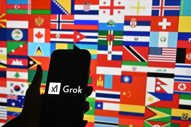 This photo illustration shows a person holding a smartphone bearing the logo of Grok, a generative artificial intelligence chatbot developed by US artificial intelligence company xAI in front of the national flags of various countries in Bengaluru on January 15, 2026. Elon Musk's platform X on January 14 announced measures to prevent its AI chatbot Grok from undressing images of real people, following global backlash over its generation of sexualized photos of women and children. India has said X removed thousands of posts and hundreds of user accounts in response to its complaints. (Photo by Idrees MOHAMMED / AFP)