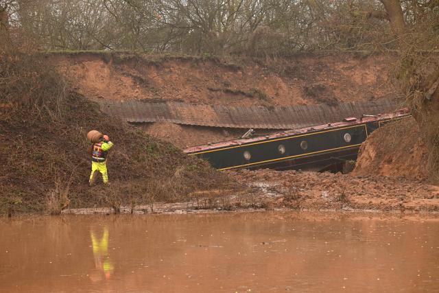 A construction worker carries hosing near a stranded canal boat, in Whitchurch, north-west England, on January 15, 2026. On December 22, 2025 Llangollen Canal collapsed, blamed on "embankment failure" by engineers, leaving narrowboat owners without their homes over Christmas. (Photo by Oli SCARFF / AFP)