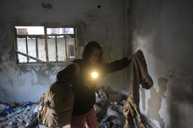 A Palestinian woman uses the torch light on her mobile phone to collect items of clothing from under the debris in a room, after an Israeli military attack on the home of the al-Houli family, in which four people were reportedly killed, west of Deir al-Balah, in the central Gaza Strip on January 16, 2026. A US-backed plan to end the war in Gaza has entered its second phase despite unresolved disputes between Israel and Hamas over alleged ceasefire violations and issues unaddressed in the first stage. (Photo by BASHAR TALEB / AFP)