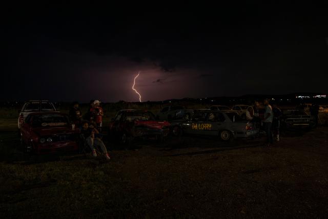 A lightning bolt strikes as members of different crews sit next to their cars at night during a round of spinning at Wheelz’n Smoke Arena in the south of Johannesburg, on January 15, 2026. Ayanda Mbele and her partner, Monde Hashe, founded the Wheelz’n Smoke Arena to create a safe space for spinners, after a lack of protections at informal spinning spots contributed to the sport’s former illegal status. Once held illegally on Thursday nights, spinning events now continue legally at the Wheelz’n Smoke Arena. (Photo by CAMILLA RICHETTI / AFP)