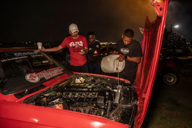 Dulaine Matthews (R) pours water on the engine of his car at the Wheelz’n Smoke Arena in south Johannesburg, on January 15, 2026. Ayanda Mbele and her partner, Monde Hashe, founded the Wheelz’n Smoke Arena to create a safe space for spinners, after a lack of protections at informal spinning spots contributed to the sport’s former illegal status. Once held illegally on Thursday nights, spinning events now continue legally at the Wheelz’n Smoke Arena. (Photo by CAMILLA RICHETTI / AFP)