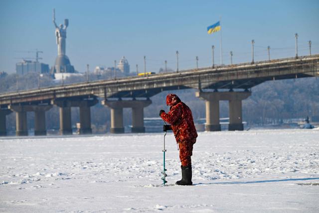 A fisherman fishes on the ice-covered Dnieper River as the Motherland Monument and a giant Ukrainian flag appear in the background in Kyiv on January 16, 2026, amid the Russian invasion of Ukraine. (Photo by Sergei GAPON / AFP)