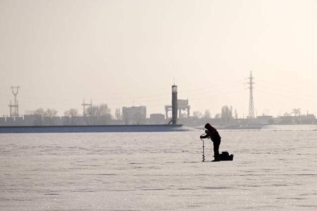 A fisherman fishes on the frozen Kyiv Reservoir located on the Dnieper north of Kyiv, also known as the Kyiv Sea, as the Kyiv Hydroelectric Station is seen in the background in Vyshgorod on January 17, 2026, amid the Russian invasion of Ukraine. (Photo by Sergei GAPON / AFP)
