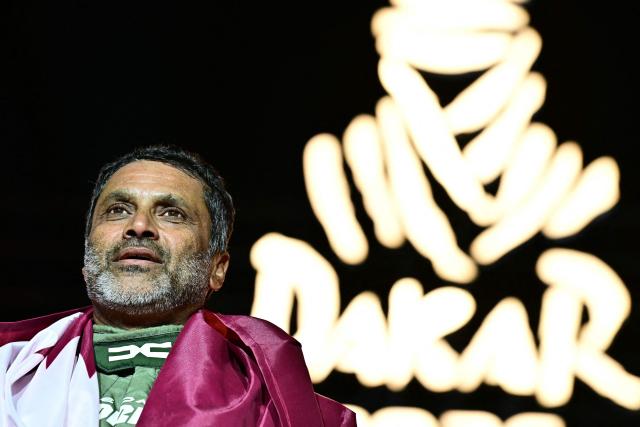 The Dacia Sandriders' Qatari driver Nasser al-Attiyah, wrapped in the national flag looks on during the trophy ceremony at the end of the 48th edition of the Dakar Rally 2026 in Yanbu, Saudi Arabia, on January 17, 2026. Qatar's Nasser Al-Attiyah secured his sixth Dakar Rally car title in Saudi Arabia. (Photo by Giuseppe CACACE / AFP)