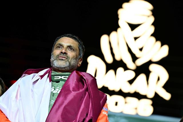 The Dacia Sandriders' Qatari driver Nasser al-Attiyah, wrapped in the national flag looks on during the trophy ceremony at the end of the 48th edition of the Dakar Rally 2026 in Yanbu, Saudi Arabia, on January 17, 2026. Qatar's Nasser Al-Attiyah secured his sixth Dakar Rally car title in Saudi Arabia. (Photo by Giuseppe CACACE / AFP)