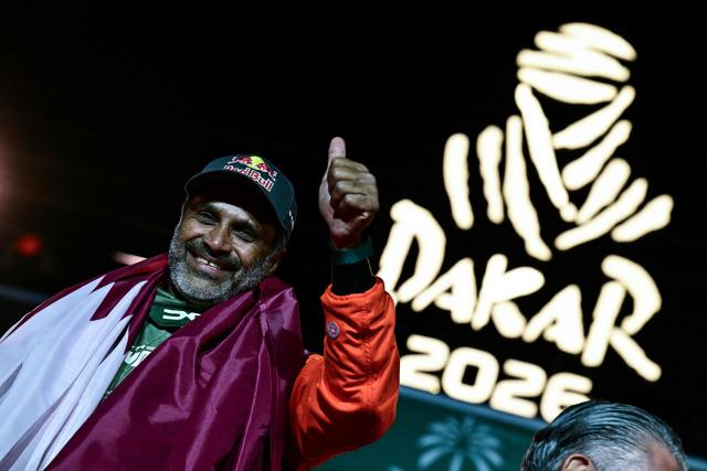 The Dacia Sandriders' Qatari driver Nasser al-Attiyah, wrapped in the national flag, gives the thumb-up during the trophy ceremony at the end of the 48th edition of the Dakar Rally 2026 in Yanbu, Saudi Arabia, on January 17, 2026. Qatar's Nasser Al-Attiyah secured his sixth Dakar Rally car title in Saudi Arabia. (Photo by Giuseppe CACACE / AFP)