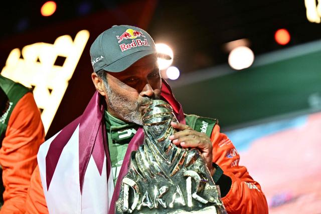 The Dacia Sandriders' Qatari driver Nasser al-Attiyah kisses the winner's trophy as he and his teammate celebrate on the podium after winning the 48th edition of the Dakar Rally 2026 in Yanbu, Saudi Arabia, on January 17, 2026. Qatar's Nasser Al-Attiyah secured his sixth Dakar Rally car title in Saudi Arabia. (Photo by Giuseppe CACACE / AFP)