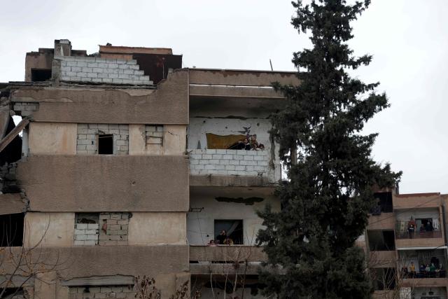 A family stands on their apartment balcony as they watch the Syrian Ministry of Defense forces drive through the streets of the northern city of Raqa, on the north bank of the Euphrates River on January 18, 2026. The Syrian President on January 18, announced an agreement with the chief of the Kurdish-led Syrian Democratic Forces that includes a ceasefire after government forces advanced in Kurdish-held areas of the north and east. The presidency published the text of the signed, 14-point deal, which includes integrating the SDF and Kurdish security forces into the defence and interior ministries, the immediate handover of Kurdish-run Deir Ezzor and Raqa provinces to the government, and for Damascus to take responsibility for Islamic State group prisoners and their families held in Kurdish-run jails and camps. (Photo by Bakr ALkasem / AFP)