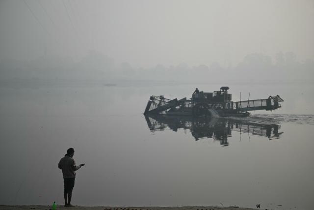 A man offers prayers as a worker collects waste using a trash skimmer machine across river Yamuna, amid dense smog in New Delhi on January 19, 2026. (Photo by Sajjad HUSSAIN / AFP)