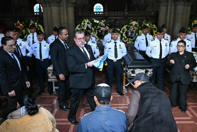 Guatemalas President Bernardo Arevalo holds a national flag in front of relatives of slain officers during a funeral ceremony at the Interior Ministry headquarters in Guatemala City on July 19, 2026. Guatemala's president on July 18 declared a 30-day nationwide state of emergency to combat criminal gangs after authorities accused the groups of killing eight police officers and holding hostages at three prisons. The killings occurred in the Guatemalan capital and surrounding areas a day after gang-affiliated inmates took 46 people hostage in the three prisons across the country to demand incarcerated gang leaders be moved to lower-security facilities. (Photo by JOHAN ORDONEZ / AFP)