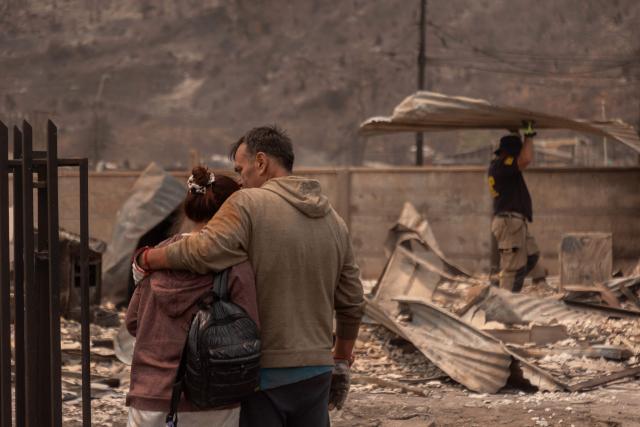 A couple embraces while a man carries debris from a burnt home in the Gabriela Mistral neighborhood after a wildfire in Lirquen, Chile, on January 19, 2026. Wildfires that have killed 19 people in southern Chile and wiped out entire towns, raged for a third day on January 19, fanned by high temperatures and strong winds at the height of the southern hemisphere summer. (Photo by Raul BRAVO / AFP)