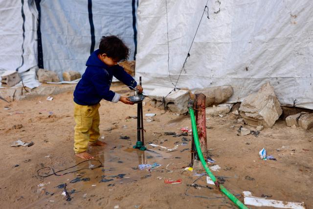 A boy fills water in a bowl next to tents in Gaza City on January 20, 2026. Day-to-day living conditions for the vast majority of Gaza residents remain extremely precarious, with more than 80 percent of infrastructure destroyed, according to the United Nations. Several humanitarian and UN workers told AFP that while the situation has improved in some areas since the ceasefire, the humanitarian response remains insufficient due to access restrictions imposed by Israeli authorities -- who deny these claims. Water and electricity networks, as well as waste management, no longer function. (Photo by Omar AL-QATTAA / AFP)