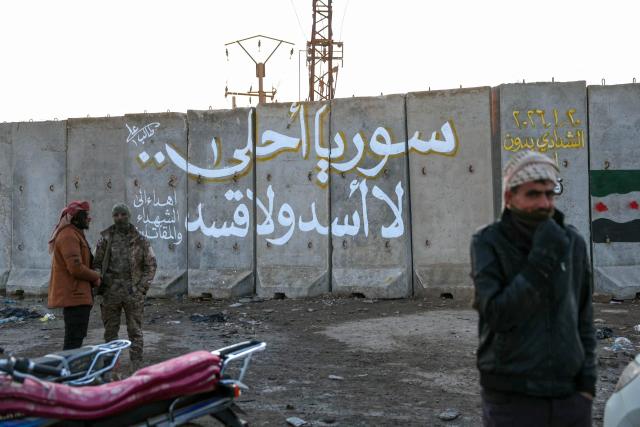 People stand by a concrete wall on which is written in Arabic “ Syria is better without Assad and Kassad” in the Syrian city of Ash Shaddadah south of the city of Hasakeh in northeast Syria on January 20, 2026. Negotiations have collapsed between the Syrian president and the chief of the country's Kurdish-led forces, a Kurdish official told AFP, as the army deployed reinforcements to flashpoint areas in the north. (Photo by OMAR HAJ KADOUR / AFP)