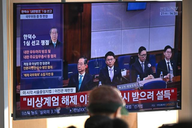 People watch a television screen showing a live broadcast of the trial verdict of South Korea's former prime minister Han Duck-soo (L on the screen) on charges related to martial law, at a train station in Seoul on January 21, 2026. A South Korean court sentenced former prime minister Han Duck-soo to 23 years in prison on January 21, for aiding and abetting a declaration of martial law that briefly suspended civilian rule. (Photo by Jung Yeon-je / AFP)