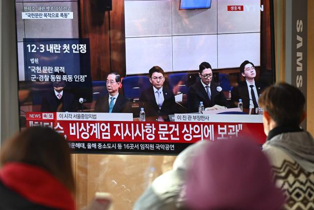 People watch a television screen showing a live broadcast of the trial verdict of South Korea's former prime minister Han Duck-soo (L on the screen) on charges related to martial law, at a train station in Seoul on January 21, 2026. A South Korean court sentenced former prime minister Han Duck-soo to 23 years in prison on January 21, for aiding and abetting a declaration of martial law that briefly suspended civilian rule. (Photo by Jung Yeon-je / AFP)