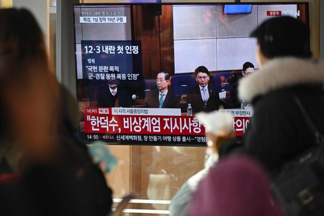 People watch a television screen showing a live broadcast of the trial verdict of South Korea's former prime minister Han Duck-soo (L on the screen) on charges related to martial law, at a train station in Seoul on January 21, 2026. A South Korean court sentenced former prime minister Han Duck-soo to 23 years in prison on January 21, for aiding and abetting a declaration of martial law that briefly suspended civilian rule. (Photo by Jung Yeon-je / AFP)